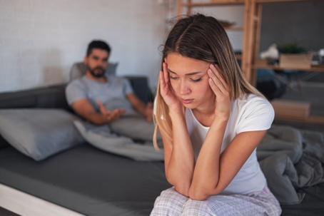 Beautiful depressed woman sitting on bed while her angry boyfriend yells at the background